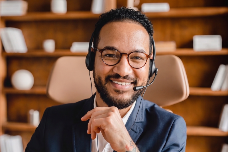 A smiling man with glasses and a headset, wearing a navy blazer over a white shirt, sitting in front of wooden bookshelves.